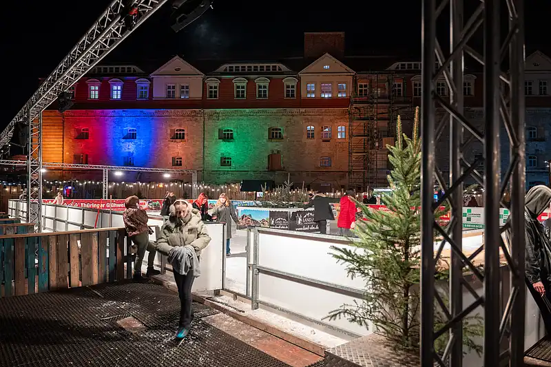 Eingang zur Eislaufbahn mit Besuchern; im Hintergrund leuchtet eine historische Fassade in Regenbogenfarben, Tanne vorn.