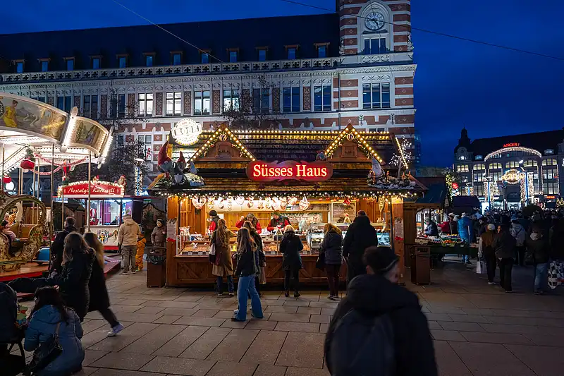 Belebter Weihnachtsmarkt auf dem Anger mit Stand „Süßes Haus“: beleuchtete Dächer und Menschen vor blau angestrahltem Gebäude.
