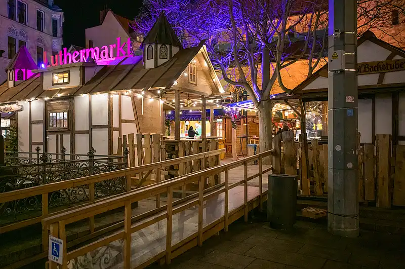 Verträumter Luthermarkt am Abend: Reihenholzstände, blau-violett leuchtender Baum und Besucher entlang des Geländers