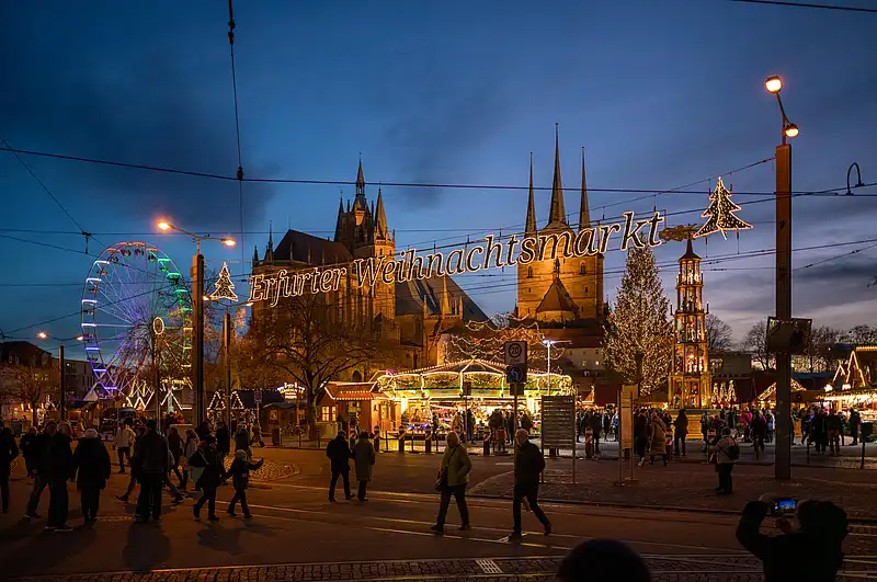 Leuchtende Schrift über der Straße, darunter strömen Besucher zum Markt; Domtürme und Riesenrad im Hintergrund.