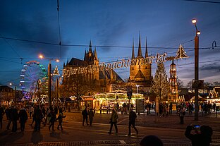 Leuchtende Schrift über der Straße, darunter strömen Besucher zum Markt; Domtürme und Riesenrad im Hintergrund.