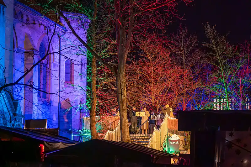 Stimmung am illuminierten Markt: Musiker und Gäste am Stand, blaue Kirchenmauer und farbige Bäume im Leuchten.