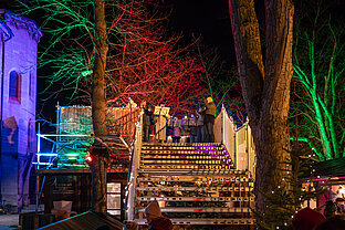 Bühnenansicht eines Konzerts am Weihnachtsmarkt: Musiker hinter Holzbarriere, umrahmt von roten und grünen Lichtbäumen.