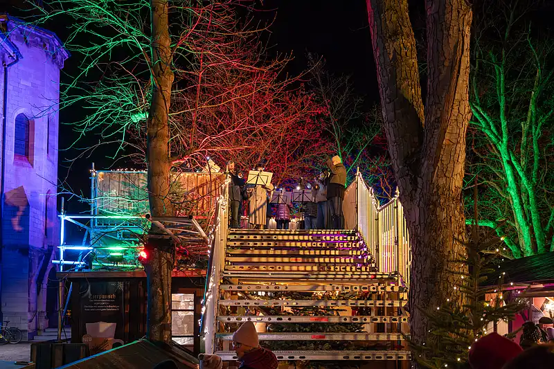 Bühnenansicht eines Konzerts am Weihnachtsmarkt: Musiker hinter Holzbarriere, umrahmt von roten und grünen Lichtbäumen.