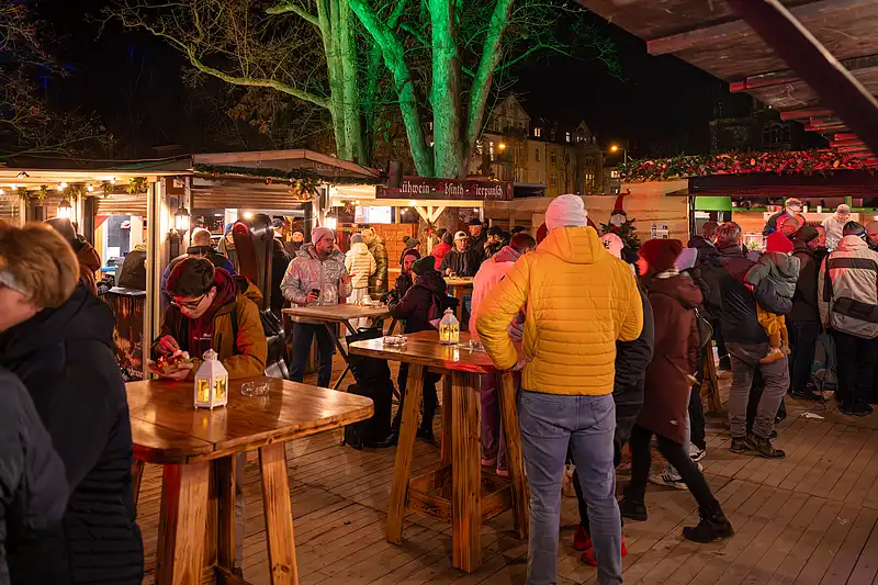 Lange Holztafeln am Weihnachtsmarkt: Gäste sitzen an beleuchteten Ständen, grün angestrahlte Bäume im Hintergrund.
