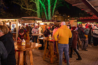 Lange Holztafeln am Weihnachtsmarkt: Gäste sitzen an beleuchteten Ständen, grün angestrahlte Bäume im Hintergrund.