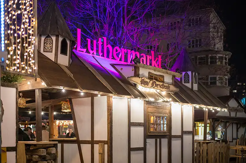 Nahaufnahme eines Fachwerkstands: über dem Dach leuchtet der Luthermarkt-Schriftzug, Lichterketten am Eingang.