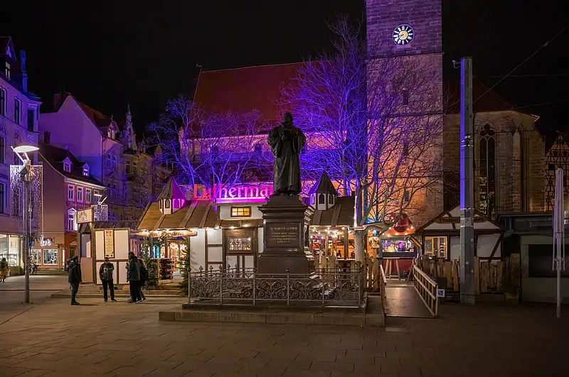 Luthermarkt-Promenade mit Luther-Denkmal im Vordergrund, warm beleuchteten Ständen und blau-violettem Baumlicht bei Nacht