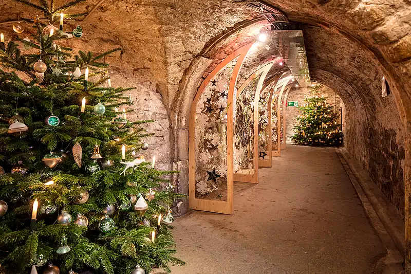 Geschmückter Weihnachtsbaum mit Kerzen und Sternenbogen in warm beleuchtetem historischem Steingewölbe.