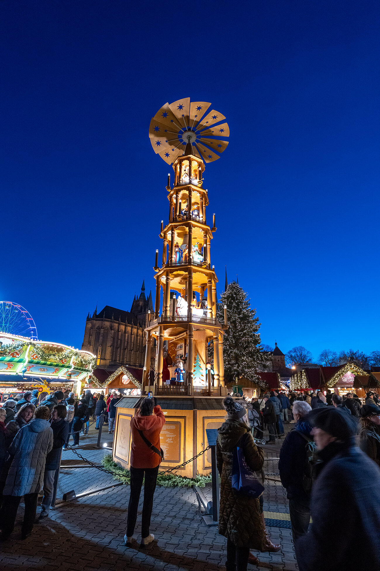 Leuchtende Weihnachtspyramide vor tiefblauem Himmel, unten stehen Besucher und fotografieren die beleuchteten Figuren.