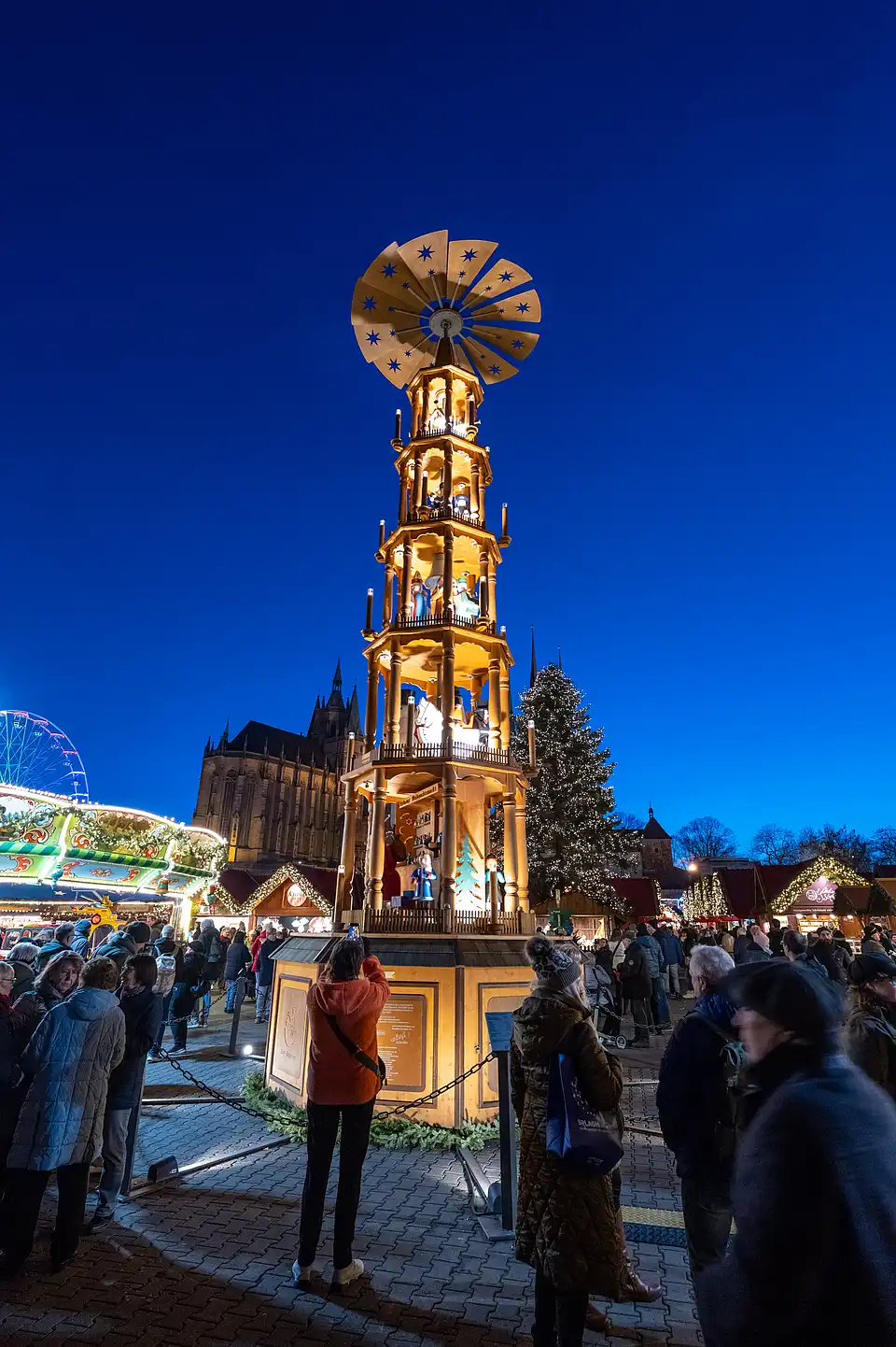 Leuchtende Weihnachtspyramide vor tiefblauem Himmel, unten stehen Besucher und fotografieren die beleuchteten Figuren.