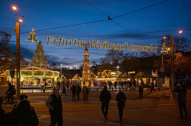 Großer leuchtender Schriftzug spannt sich über den Platz, darunter Buden, Karussell und Spaziergänger in der Nacht.