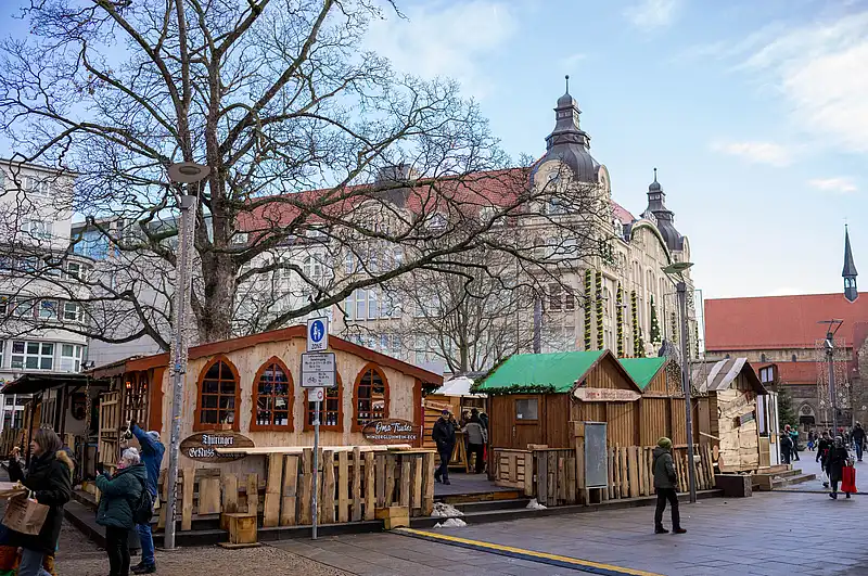 Vor einer Kaufmannskirche verteilen sich Holzbuden auf dem Platz, Passanten schlendern zwischen kahlen Bäumen