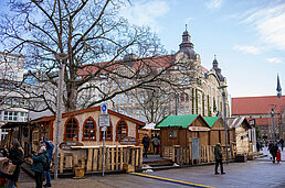 Vor einer Kaufmannskirche verteilen sich Holzbuden auf dem Platz, Passanten schlendern zwischen kahlen Bäumen