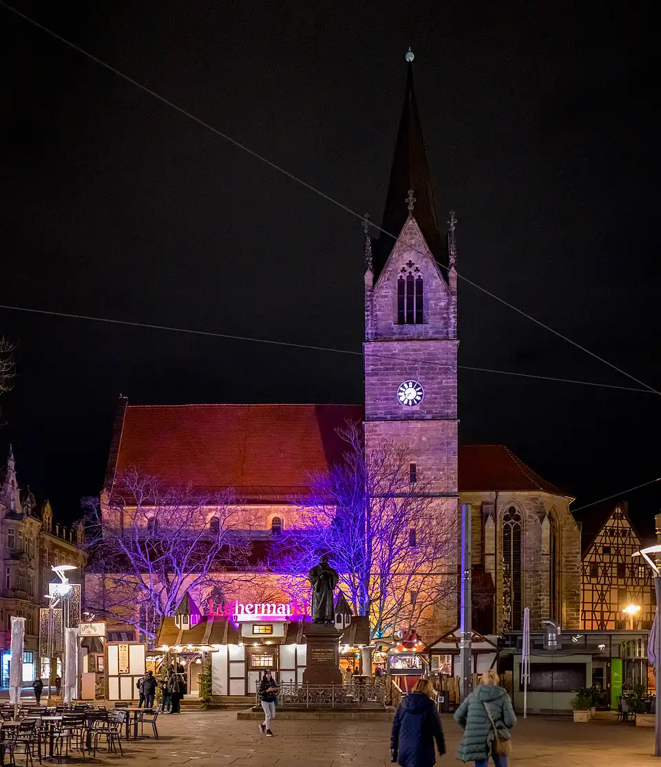 Luthermarkt bei Nacht: beleuchteter Kirchturm mit lila Licht, vor dem Platz Buden und Spaziergänger in Mänteln