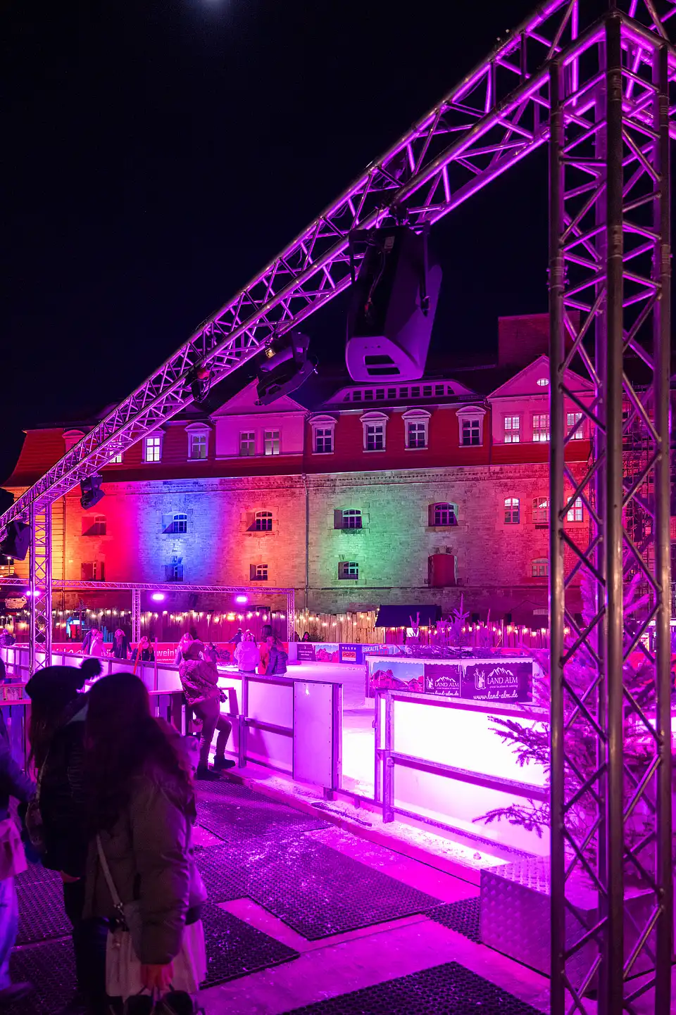 Violett leuchtende Traverse über der Eisbahn, dahinter bunt angestrahlte Fassade und Besucher am Rand.