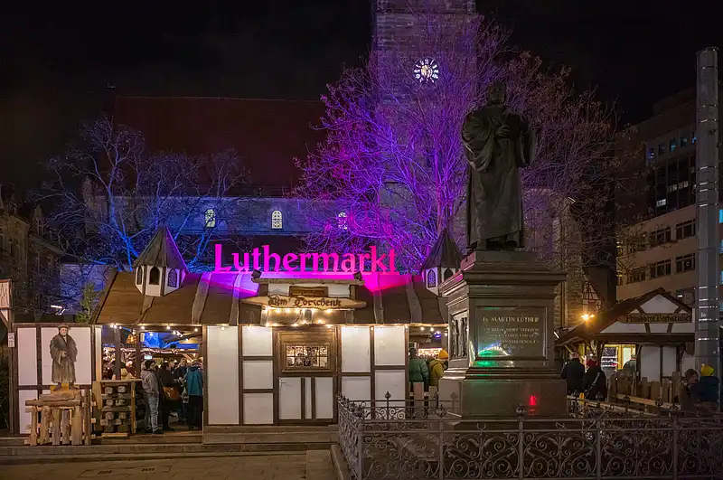 Eingang zum Luthermarkt bei Nacht, Fachwerkbuden und Lutherdenkmal vor violett beleuchteter Kirche.