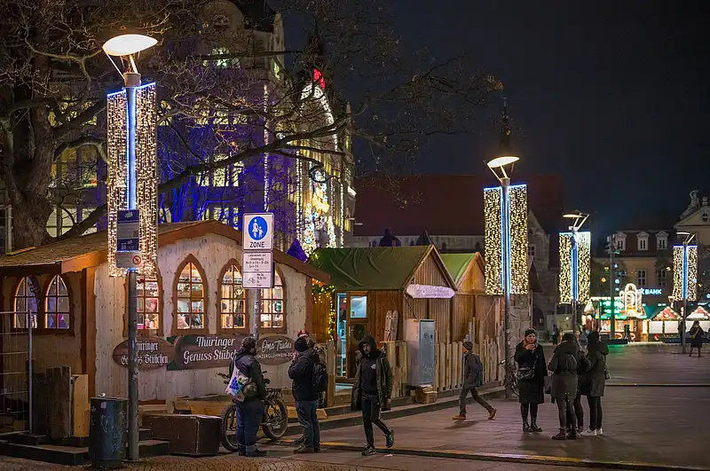 Reihe von Holzhütten am Weihnachtsmarkt, Passanten unter hohen Lampen mit Lichtervorhängen in der Nacht.