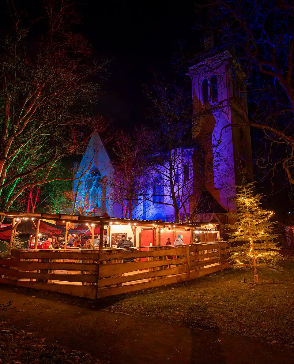 Wintermarkt am Abend: blau und violett beleuchteter Glockenturm, davor Lichterreihen und warme Verkaufsstände.