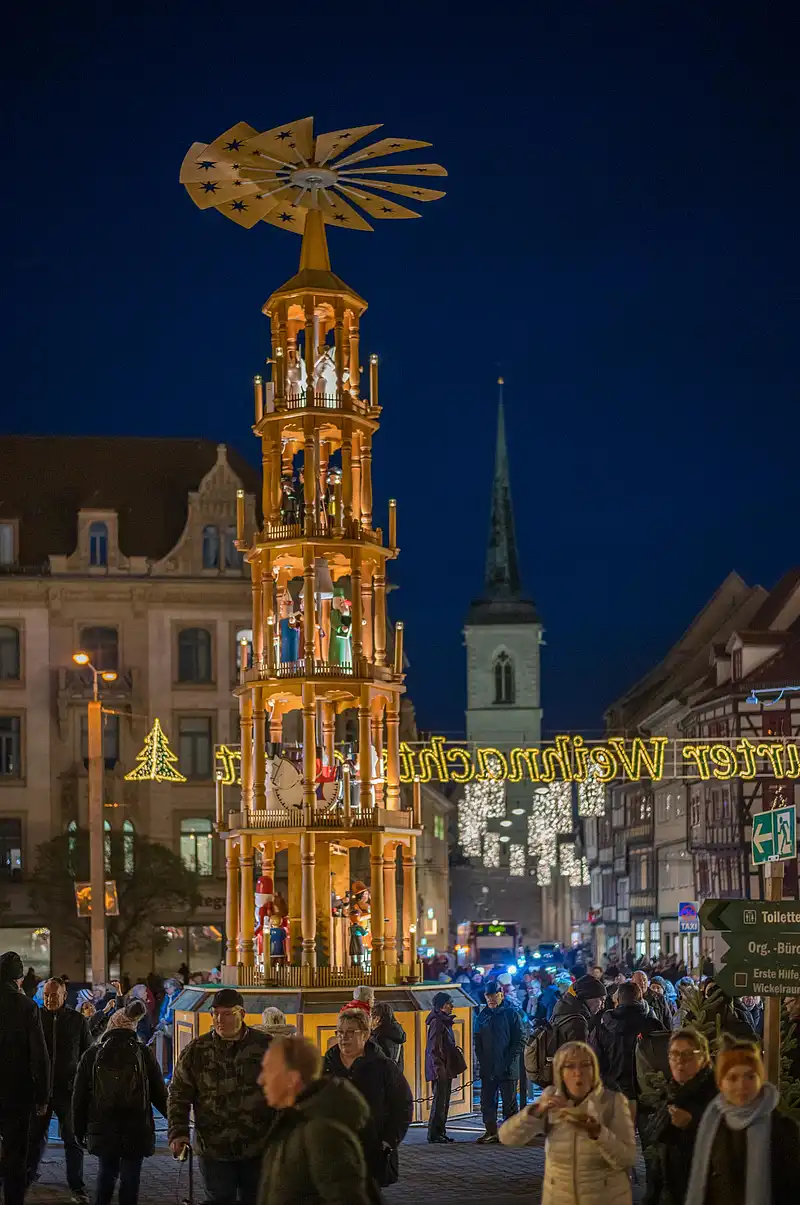 Hohe Weihnachtspyramide mit Figuren und Flügelrad steht auf der Straße, umgeben von Passanten und Lichterdeko.