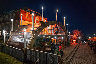 Seitansicht des Markt-Pavillons: Gäste am Eingang, Lichterketten und orange angestrahlter Baum entlang des Weges.