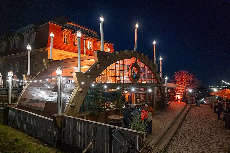 Seitansicht des Markt-Pavillons: Gäste am Eingang, Lichterketten und orange angestrahlter Baum entlang des Weges.