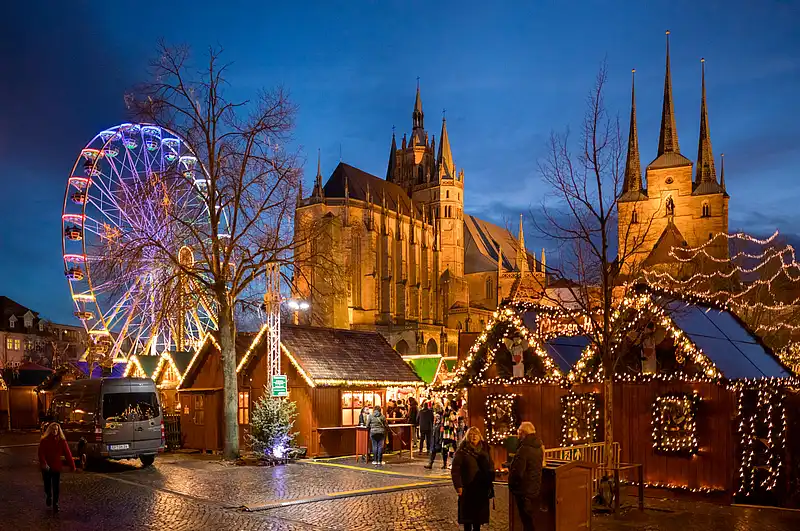 Beleuchtetes Riesenrad neben dem Dom, davor festlich geschmückte Marktbuden und Besucher im Abendblau.