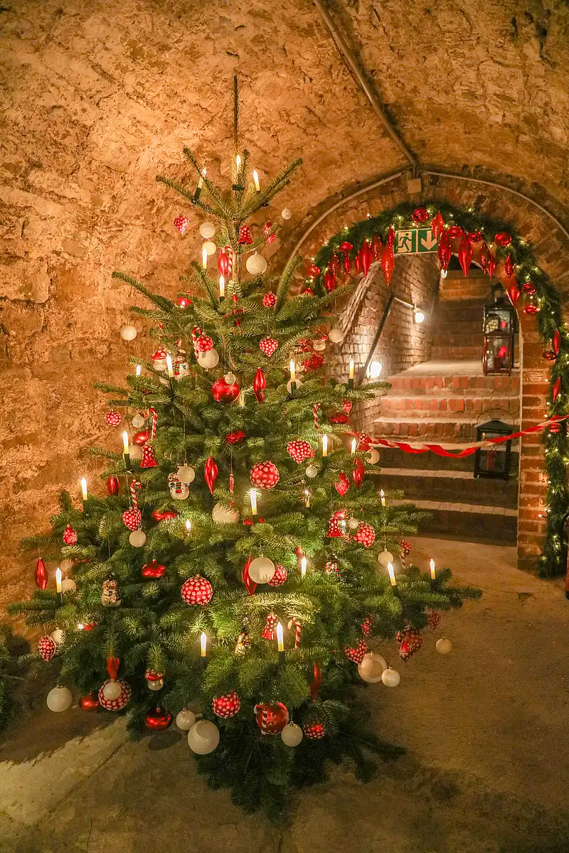 Rot-weiß geschmückter Weihnachtsbaum mit Kerzenlichtern in steinerner Gewölbehalle vor Girlandenbogen.