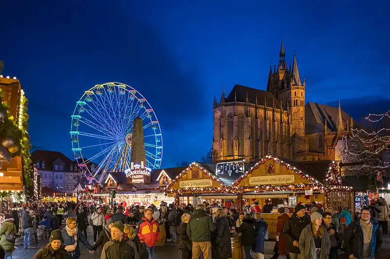 Große Menschenmenge auf dem Weihnachtsmarkt, dahinter Dom und ein farbig beleuchtetes Riesenrad am Platz.