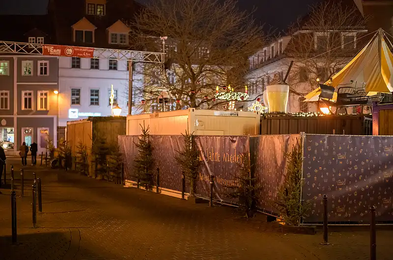 Ruhiger Weihnachtsmarktweg bei Nacht mit Bannern, kleinen Tannen und warmen Lichtlinien entlang der Zäune