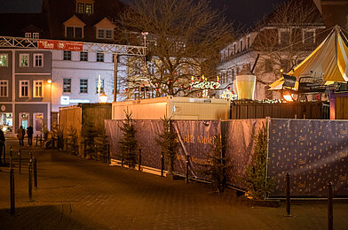 Ruhiger Weihnachtsmarktweg bei Nacht mit Bannern, kleinen Tannen und warmen Lichtlinien entlang der Zäune