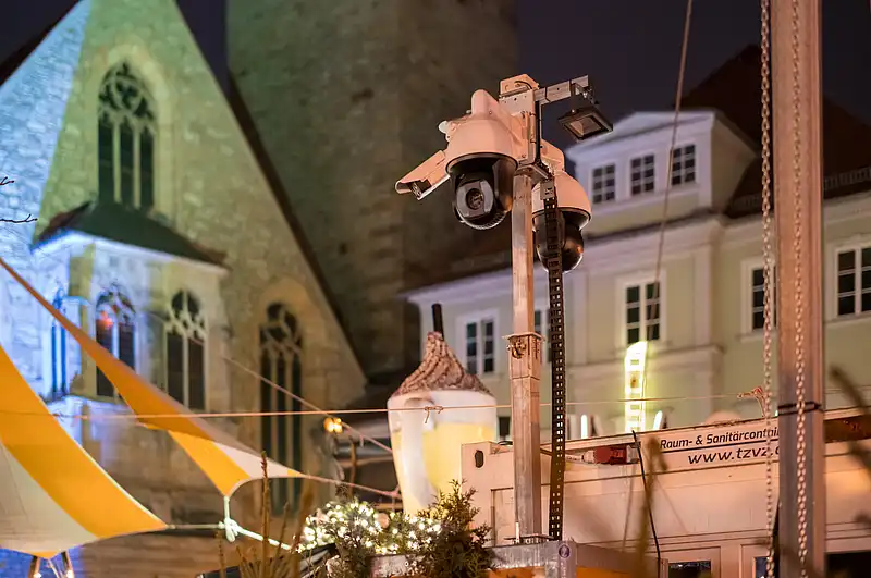 Nahaufnahme Überwachungskameras am Weihnachtsmarkt, Scheinwerfer über Tannen und gestreiften Zelten vor Fassade