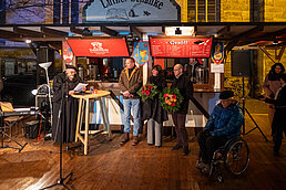 Stand mit rotem Schild, Menschen im Gespräch, Blumen auf dem Tresen und Scheinwerfer im Hintergrund