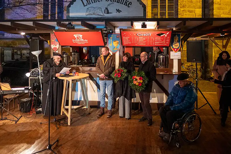 Stand mit rotem Schild, Menschen im Gespräch, Blumen auf dem Tresen und Scheinwerfer im Hintergrund
