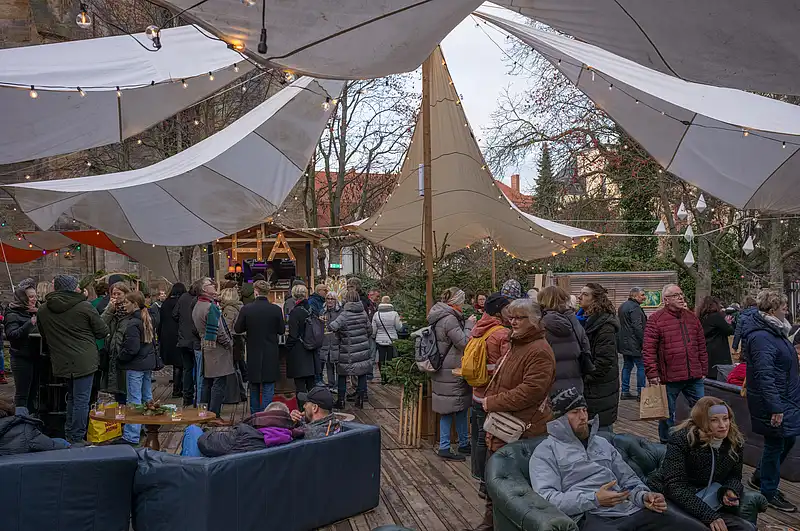 Weihnachtsmarkt unter Zeltdächern: winterliche Besucher am Tisch, offene Budenflucht und warmes Licht zwischen Bäumen