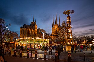 Festliche Szene mit Weihnachtspyramide, Karussell und vielen Besuchern, dahinter die Türme der Kirchen im Abendlicht.