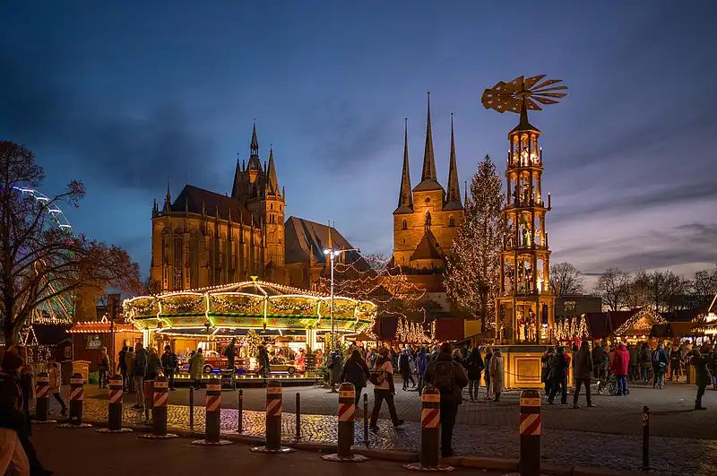 Festliche Szene mit Weihnachtspyramide, Karussell und vielen Besuchern, dahinter die Türme der Kirchen im Abendlicht.
