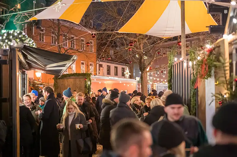Gedrängter Weihnachtsmarktweg bei Nacht mit gelb-weißen Zelten, Lichterketten und rundem Lichterdekor über dem Publikum