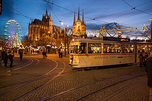Straßenbahn fährt an geschwungenen Gleisen vorbei, darüber leuchtet der Weihnachtsmarkt-Schriftzug vor Domkulisse.