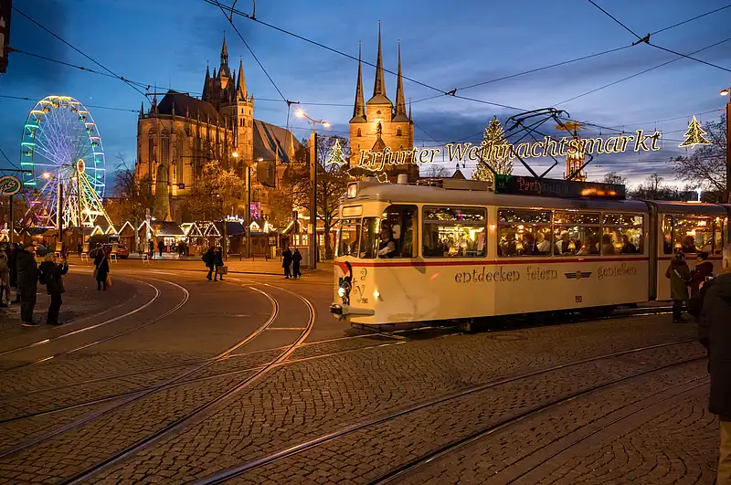 Straßenbahn fährt an geschwungenen Gleisen vorbei, darüber leuchtet der Weihnachtsmarkt-Schriftzug vor Domkulisse.